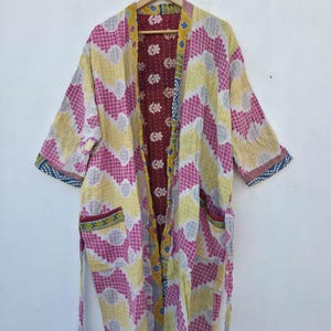 Könnte beinhalten: Ein farbenfroher Kimono-Mantel mit einem geometrischen Muster in Rosa, Gelb und Weiß. Der Mantel hat ein weinrotes Innenfutter mit Blumenmustern und hängt an einem Holzbügel. Er hat Taschen und einen Bindegürtel.