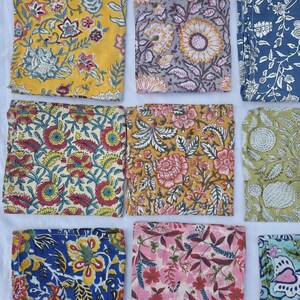 May include: A collection of colorful, square fabric napkins with floral patterns. The napkins feature various designs, including sunflowers, roses, and other botanical motifs in shades of yellow, blue, pink, and green. The fabric appears to be cotton or linen.