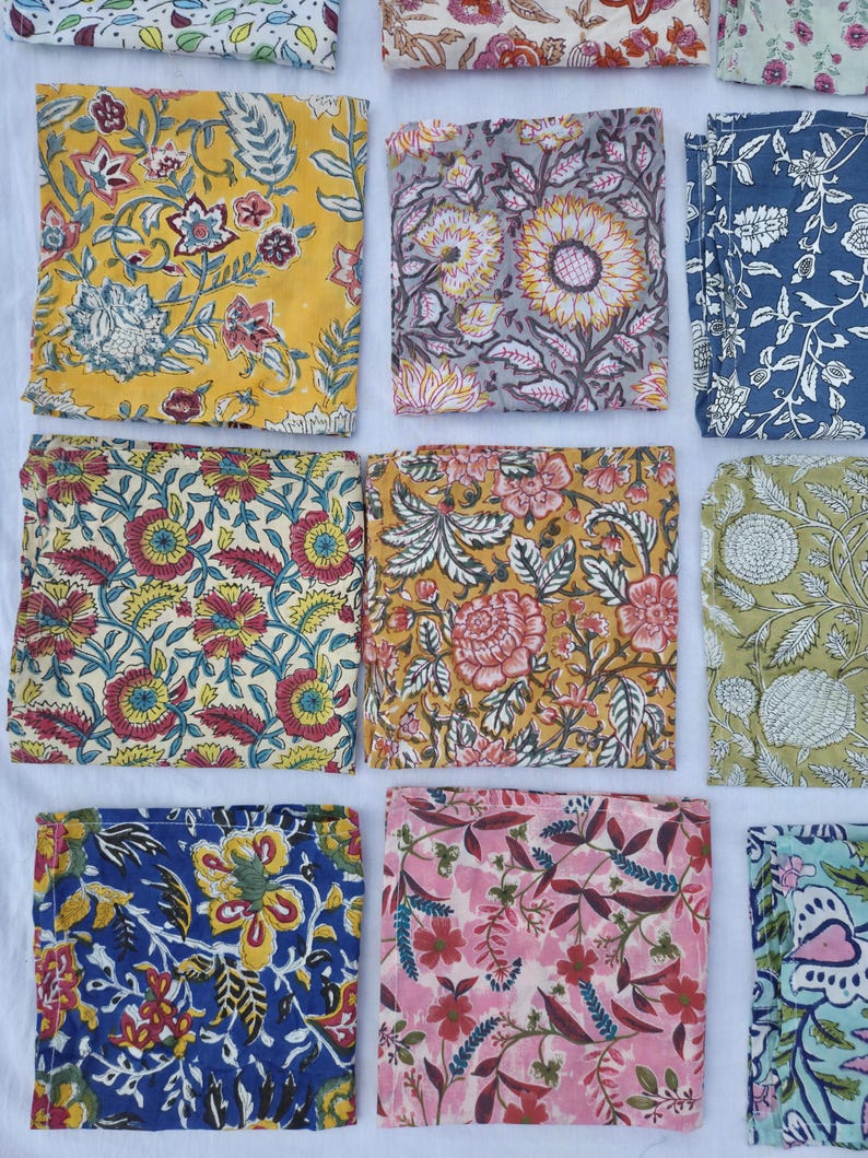 May include: A collection of colorful, square fabric napkins with floral patterns. The napkins feature various designs, including sunflowers, roses, and other botanical motifs in shades of yellow, blue, pink, and green. The fabric appears to be cotton or linen.