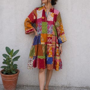 May include: A patchwork dress in a variety of colors, including red, orange, pink, green, and blue. The dress has long sleeves and a tiered skirt. The model is wearing brown sandals. A potted plant is to the left of the model.