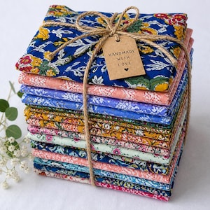 May include: A stack of colorful fabric squares with floral patterns, tied with twine and a tag that reads "Handmade with Love". The fabrics feature shades of blue, pink, yellow, and green, creating a vibrant and artistic display.