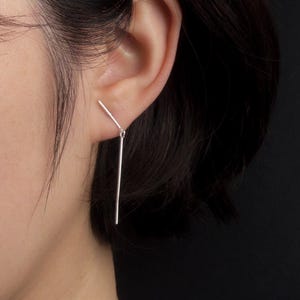 May include: A close-up of a silver bar earring. The earring is a simple, modern design with a straight bar hanging from a smaller bar that sits in the ear. The background is black.