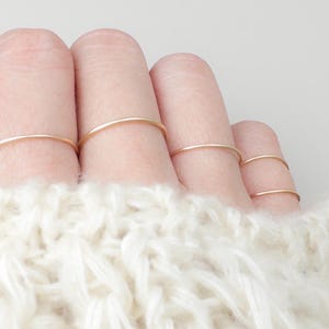 May include: A hand adorned with several delicate, gold-coloured rings. The rings are plain bands, and the hand is partially obscured by a white, textured knitted jumper. The rings are on the index, middle, ring, and little fingers.