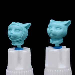 May include: Two light blue cat head figurines on white bases. One cat head wears a helmet, the other does not. The figurines are attached to white, cylindrical objects with blue rings. The background is black.