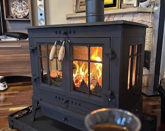Puede incluir: Una estufa de leña negra y rectangular con chimenea. La estufa tiene puertas de vidrio que revelan un fuego encendido. La estufa tiene una bandeja decorativa en la parte inferior. La estufa está en una habitación con chimenea de piedra.