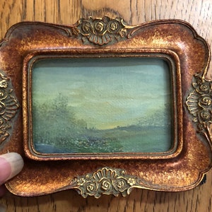 May include: A small framed painting of a landscape scene with a blue sky, green grass, and trees. The frame is gold with a decorative design.