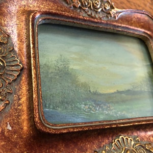May include: A small, framed painting of a landscape scene with a blue sky, green trees, and a field in the foreground. The frame is gold and has a decorative design.