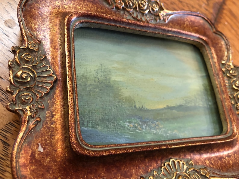 May include: A small, framed painting of a landscape scene with a blue sky, green trees, and a field in the foreground. The frame is gold and has a decorative design.
