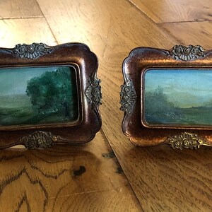 May include: Two small, ornate, gold-toned picture frames with glass fronts, each displaying a landscape painting. The painting on the left features a green tree in a field, while the painting on the right shows a field with a blue sky and a few trees.