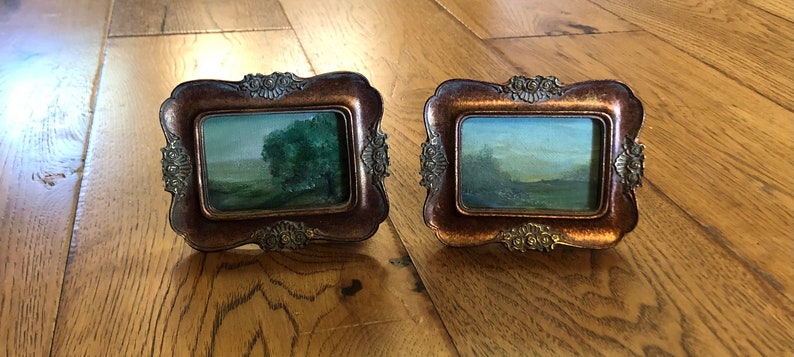 May include: Two small, ornate, gold-toned picture frames with glass fronts, each displaying a landscape painting. The painting on the left features a green tree in a field, while the painting on the right shows a field with a blue sky and a few trees.