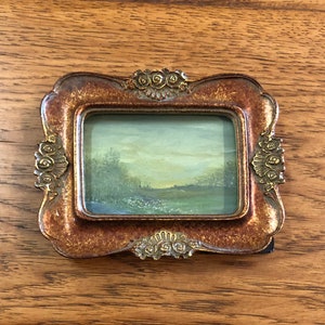 May include: A small, framed landscape painting with a gold-colored frame. The painting depicts a green field with trees in the background and a cloudy sky.