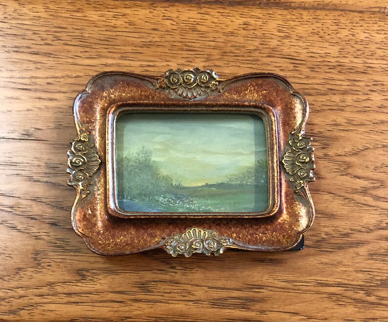 May include: A small, framed landscape painting with a gold-colored frame. The painting depicts a green field with trees in the background and a cloudy sky.