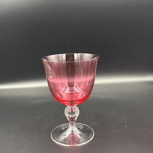 May include: A cranberry red glass goblet featuring a rounded bowl with vertical panel cuts. The stem includes a clear glass spherical knop, and the base is a circular clear glass foot. The glass transitions from a deep red bowl to a clear stem.