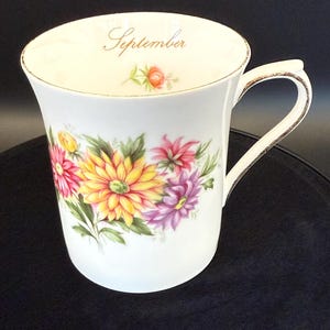 May include: White ceramic mug with a floral design featuring yellow, pink, and purple flowers. The word "September" is written in gold script near the rim. The mug has a gold trim on the rim and handle.
