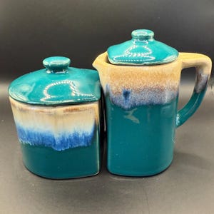 May include: A teal ceramic creamer and sugar bowl set with matching lids. The creamer has a handle and a spout. The sugar bowl is square. Both pieces have a gradient design with brown, white, and blue accents.