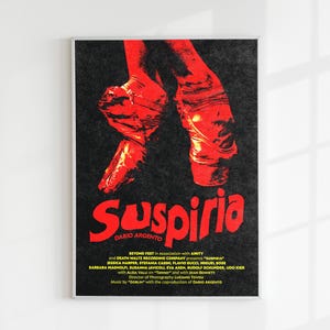 May include: A framed poster featuring the word "Suspiria" in red, stylized lettering. The poster displays a pair of red ballet shoes and text at the bottom. The background is black.