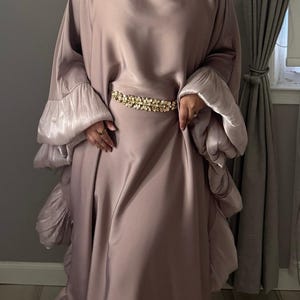 May include: A mauve-colored satin dress with long, ruffled sleeves and a matching head covering. The dress is cinched at the waist with a gold-colored, jeweled belt. The dress has a flowing, elegant design.
