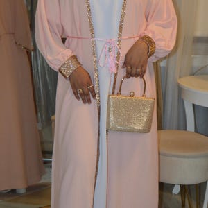 May include: A light pink open-front robe with gold sequin trim and cuffs. The robe is worn over a white dress and cinched with a pink belt. A gold glitter handbag is held in front.