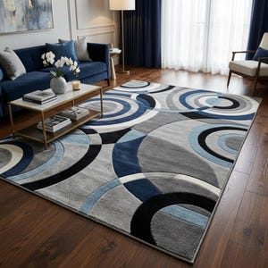 May include: A patterned rug with a circular design in shades of grey, blue and black. The rug is placed on a dark wooden floor. The design creates a modern, abstract pattern, suitable for a living room or similar space.