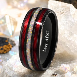 May include: A black ring featuring a central inlay of iridescent crushed opal flanked by two deep red grooved bands. The interior of the band is engraved with the text Ever After in a clean white font, contrasting against the dark metal finish.