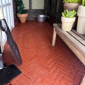 May include: A miniature garden scene with a red brick floor laid in a herringbone pattern. The scene includes terracotta pots with plants, a wooden bench, and a metal basin. A gardening tool rests on the floor.