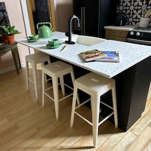May include: Miniature kitchen island with a white countertop, black base, and three white stools. A green teapot and teacups sit on the counter. A black faucet and sink are visible. A magazine is on the counter.