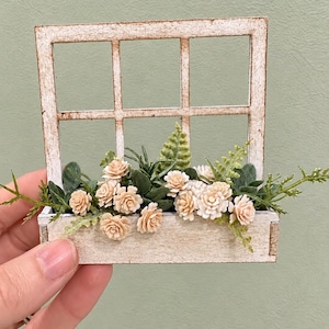 May include: A miniature wooden window frame with a rectangular planter box attached at the base. The frame and box feature a distressed white finish. The planter is filled with small, light peach-colored artificial flowers and green foliage.
