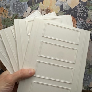 May include: A stack of off-white rectangular panels with a raised rectangular design. The panels have a vertical grooved pattern on the sides. The background features a floral wallpaper with blue, yellow, and orange flowers.