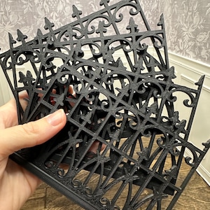 May include: A stack of black, decorative miniature fences with ornate scrollwork and pointed finials. The fences have a repeating pattern of vertical and diagonal bars, creating a detailed, gothic-inspired design. The fences are likely for a dollhouse or miniature scene.