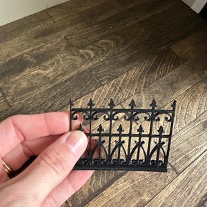 Puede incluir: Una pequeña valla decorativa negra con adornos de volutas y pináculos puntiagudos. La valla se sostiene sobre un fondo de suelo de madera. Este artículo es probablemente para una casa de muñecas o una escena en miniatura.