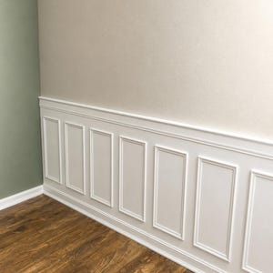 May include: White wainscoting wall paneling featuring a series of rectangular frames with raised trim molding. The paneling is installed along the lower portion of a wall, topped with a horizontal chair rail, and set against a beige upper wall.