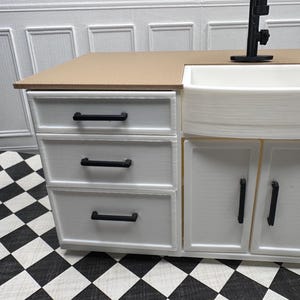 Könnte beinhalten: Ein Miniatur-Küchenschrank-Set in Weiß mit einer beigefarbenen Arbeitsplatte, einem weißen Bauernhaus-Waschbecken und schwarzen Beschlägen. Der Schrank hat drei Schubladen und zwei Türen. Der Boden hat ein schwarz-weißes Schachbrettmuster.