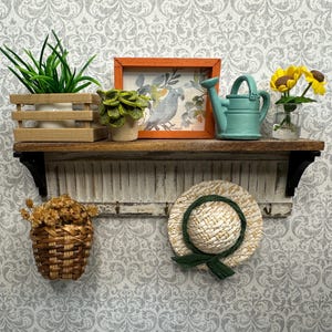 Op de afbeelding: Een miniatuur decoratief plankje met diverse items. Het heeft een kleine houten kist met een groene plant, een ingelijste foto, een blauwe gieter en een vaas met gele bloemen. Daaronder een gevlochten mand en een strohoed met een groen lint.