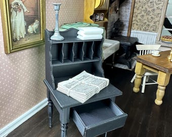 May include: A miniature dark gray wooden secretary desk with a pull-out drawer, a top shelf featuring a silver candlestick and a stack of white books, and a flat surface holding a bundle of miniature newspapers tied with white string.