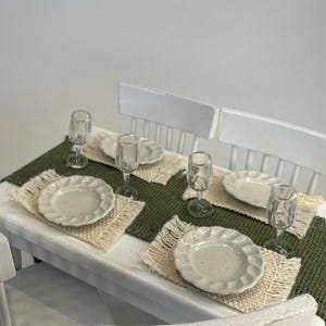 May include: A miniature dining table set for four. The table is white with a green runner. Each place setting includes a white plate, a clear wine glass, and a woven placemat. The chairs are white.