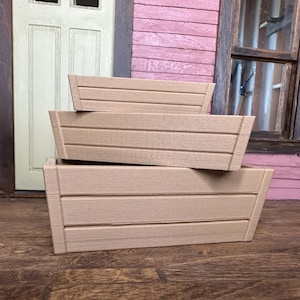 Peut inclure: Trois jardinières rectangulaires en bois de couleur marron clair, empilées les unes sur les autres. Chaque jardinière présente des rainures horizontales et une forme trapézoïdale. En arrière-plan, une porte et une fenêtre.