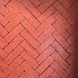 May include: Close-up of a red brick pattern arranged in a herringbone design. The bricks have a textured surface and are slightly glossy. The pattern is a repeating zig-zag, creating a visually interesting surface.