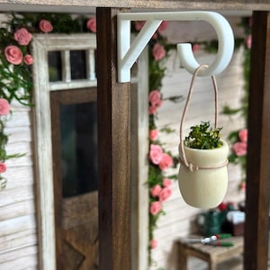 May include: A miniature dollhouse scene featuring a white bracket holding a small, cream-colored pot with greenery. The pot hangs by a thin, tan string. Pink roses and greenery decorate the background.