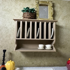 May include: A miniature kitchen scene featuring a beige plate rack with white plates, a gold-framed mirror, and a small potted plant. Two teacups and saucers sit on a shelf below. A black candlestick and a yellow pumpkin are in the foreground.