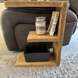 May include: A three-tiered, wooden side table in a rustic style. A glass jar candle and a book titled "SAS" sit on the top shelf. The middle shelf features a black storage bin. The table is made of light brown wood.