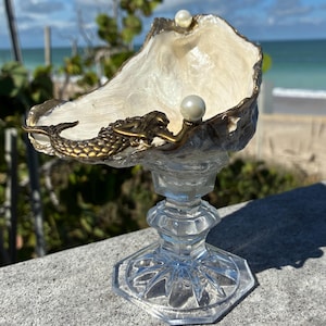 May include: A decorative seashell bowl with a gold-toned mermaid accent, holding two pearls. The shell sits atop a clear, faceted glass pedestal. The background features a beach scene with blue sky and ocean.