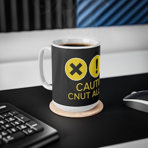 May include: A white ceramic coffee mug with a black exterior featuring yellow caution symbols and text. The mug sits on a cork coaster, filled with coffee, and is placed on a black surface.