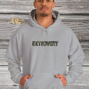 Może przedstawiać: Jasnoszara bluza z kapturem z napisem "EXTROVERT" w kamuflażu na piersi. Bluza z kapturem ma kaptur ze sznurkiem i przednią kieszeń. Model ma na sobie bluzę z kapturem.