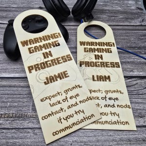 May include: Two wooden door hangers with a hole at the top, featuring the text "WARNING! GAMING IN PROGRESS" and personalized names. One says "Jamie" and the other "Liam". Below, text reads "Expect grunts, lack of eye contact, and nods if you try communication."