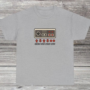 May include: A light gray t-shirt featuring a retro video game controller design in brown, beige, and red. Below the controller is the text "KNOW YOUR CHEAT CODE" in red. The shirt is made of a soft material.