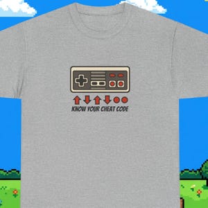 Może przedstawiać: Szary melanżowy t-shirt z grafiką kontrolera do gier w stylu retro nad sekwencją czerwonych strzałek kierunkowych i kółek, z napisem "KNOW YOUR CHEAT CODE" wydrukowanym czarnymi literami pod ilustracją kontrolera.