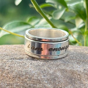 May include: A silver-colored spinner ring with the inscription "I am enough" engraved on the band. The ring is set against a natural stone surface, with green foliage in the background.