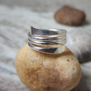 May include: A silver ring with a spiral design. The ring is made up of several bands that coil around the finger. The ring is placed on a light brown stone, with other stones in the background.
