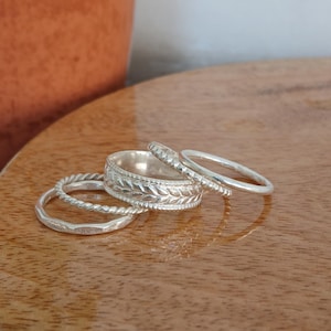 May include: Four silver rings of varying designs are arranged on a wooden surface. One ring has a twisted rope design, another has a leaf pattern, and two are simple bands. The rings are displayed in a close-up shot.