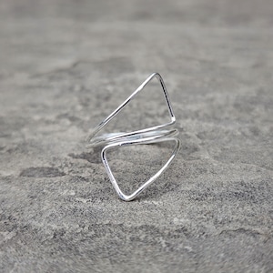 Puede incluir: Un anillo de plata con un diseño moderno. El anillo presenta dos triángulos abiertos, uno encima del otro, conectados por una banda que rodea el dedo. El anillo está hecho de un metal brillante y reflectante.